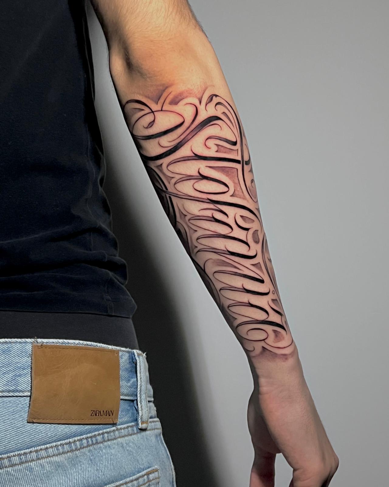 Lettering ornamentale che copre l’avambraccio, linee nere e ombreggiature grigie, tatuato da Armando Sideri